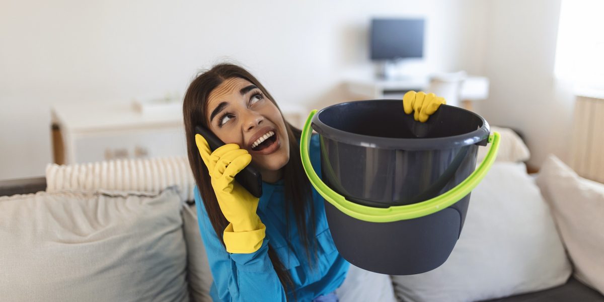 Personne assise sur un canapé tenant un seau sous une fuite au plafond, téléphone à l'oreille