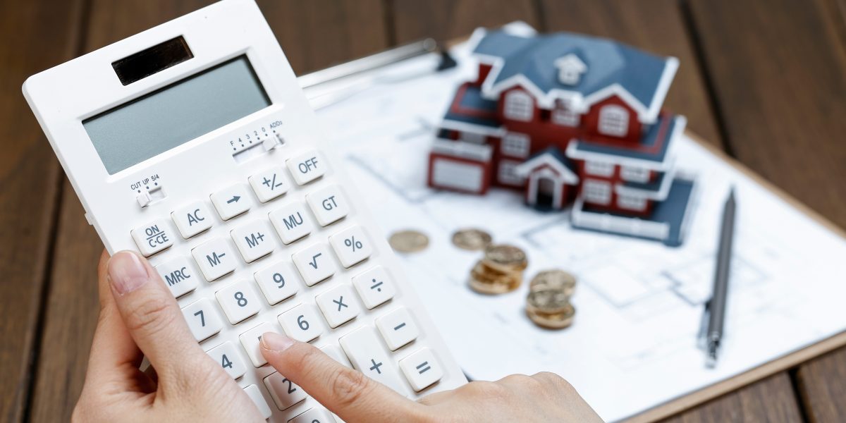 Main tapant sur une calculatrice, maquette de maison et pièces de monnaie à côté